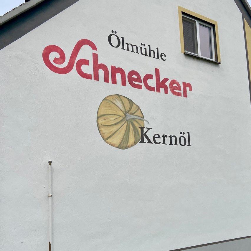 A white building with a red logo for Schneckenkernöl, featuring a pumpkin icon, painted on its wall.