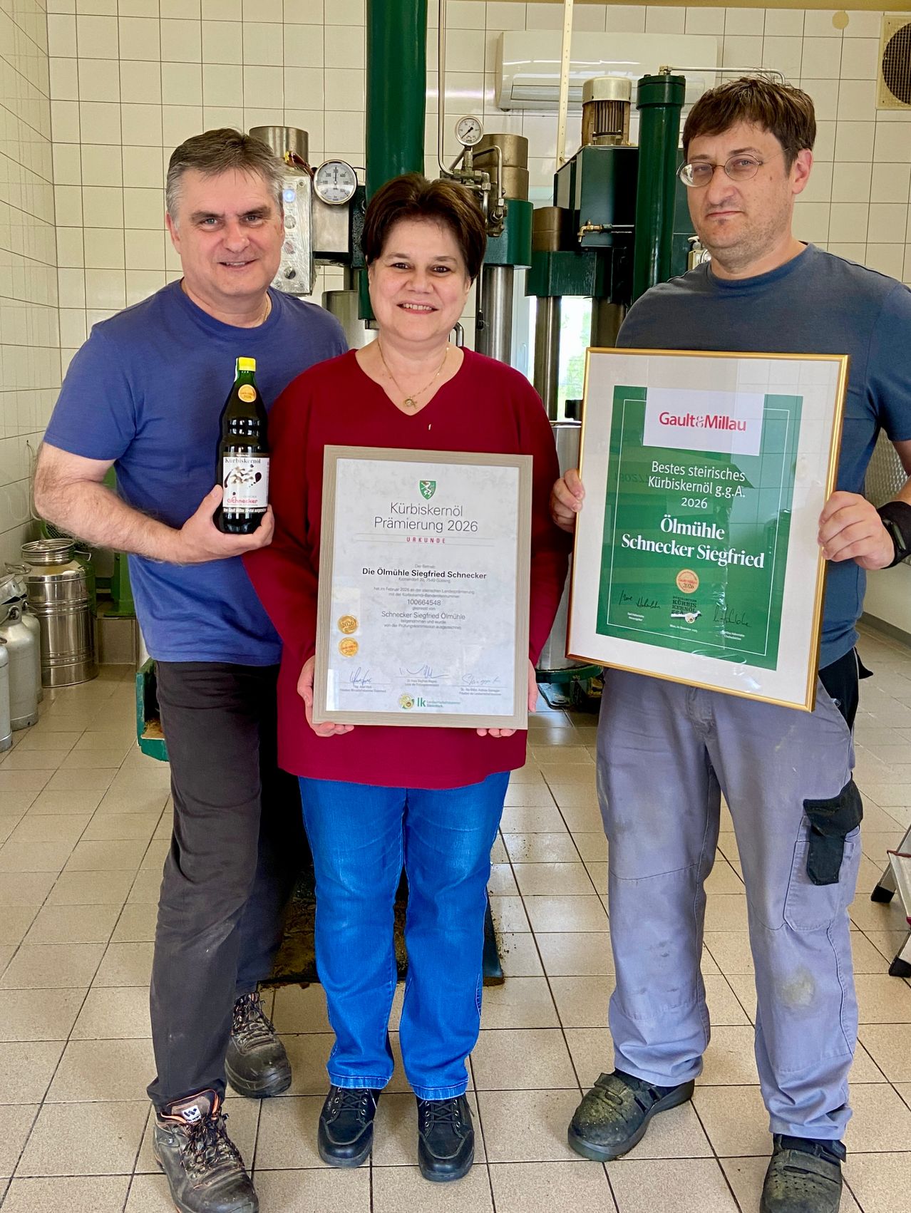 Three people stand in a factory room. A woman holds a certificate and a bottle of green liquid. The men on either side hold framed certificates. Behind them are metal containers.