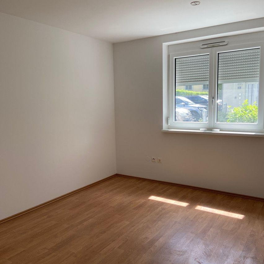 Ein leerer Raum mit Holzböden und weißen Wänden. Das Zimmer hat ein Fenster mit weißen Jalousien und Blick auf geparkte Autos draußen.