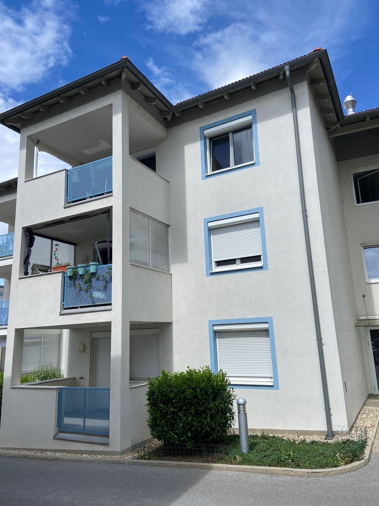 Ein weißer Apartmentkomplex mit blauer Fenster- und Balkonumrandung. Er hat drei Stockwerke und mehrere Balkone mit Metallgeländern.
