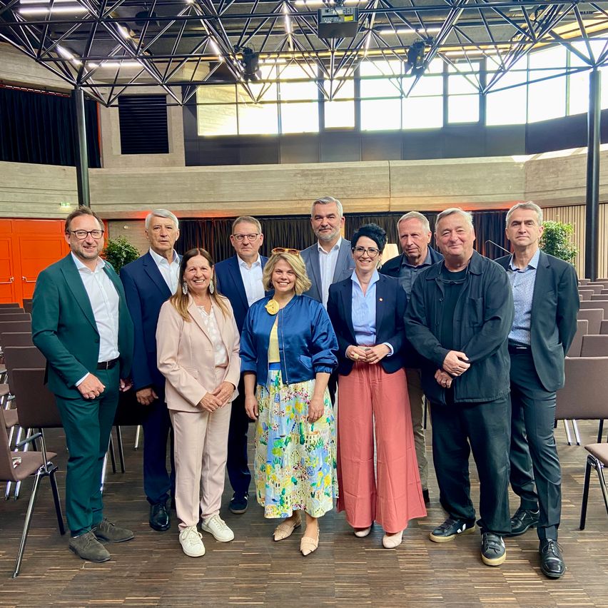 Eine Gruppe von Erwachsenen steht in einem Konferenzraum mit moderner Architektur. Sie lächeln und schauen in die Kamera. Sie tragen formelle Kleidung.