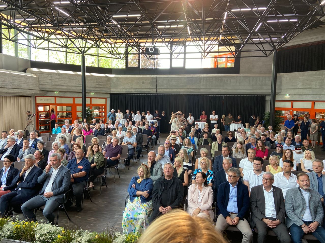 Eine Menschenmenge sitzt in Reihen in einem modernen Gebäude mit hohen Decken. Einige tragen Brillen und einige haben Namensschilder. Der Raum ist mit natürlichem Licht durch große Fenster gefüllt.