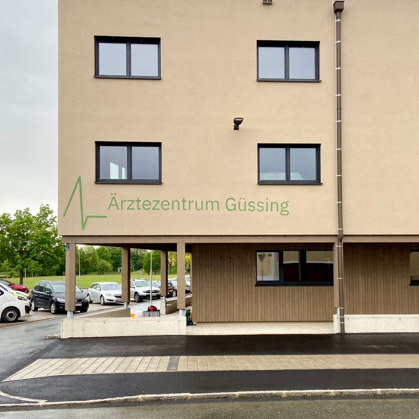 Das Arztezentrum Gussing Gebäude ist eine medizinische Einrichtung mit mehreren Fenstern. Autos sind in der Nähe geparkt, und das Gebiet ist von Bäumen umgeben.