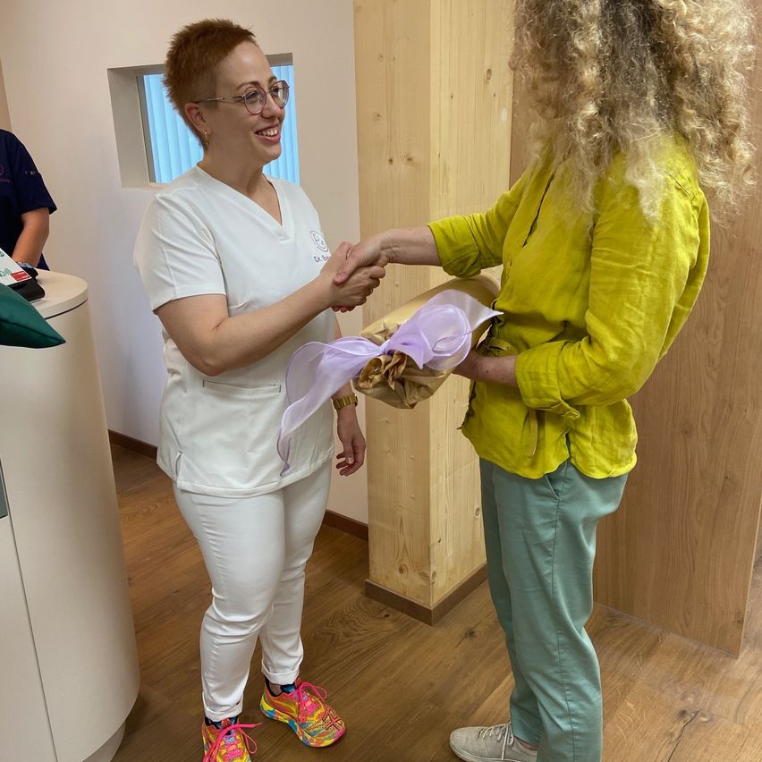 Zwei Frauen schütteln sich in einem Raum die Hände. Eine Frau ist in einem weißen Outfit gekleidet und die andere in einem gelben Hemd und grüner Hose. Sie halten ein Geschenk in braunes Papier gewickelt.