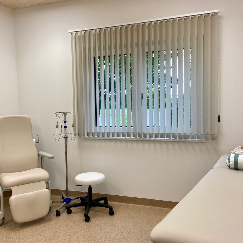 Ein Zimmer mit einer weißen Wand, einem Bett, einem Rollstuhl, einem Hocker und einem medizinischen Ständer vor einem Fenster mit Jalousien.
