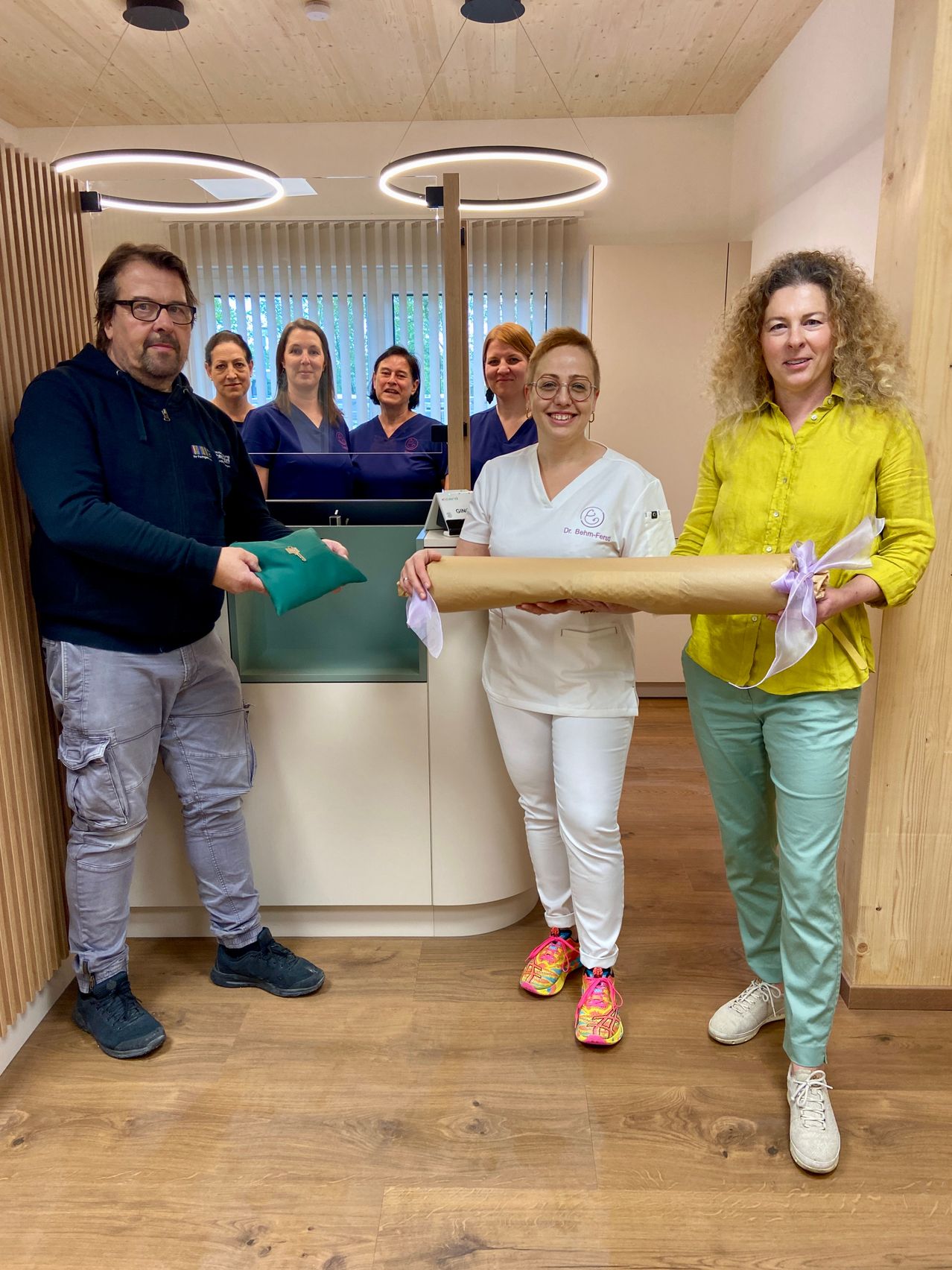 Eine Gruppe von Menschen in einer medizinischen Umgebung, ein Mann und zwei Frauen halten ein gerolltes Stück Papier. Dahinter sind andere medizinische Mitarbeiter zu sehen.