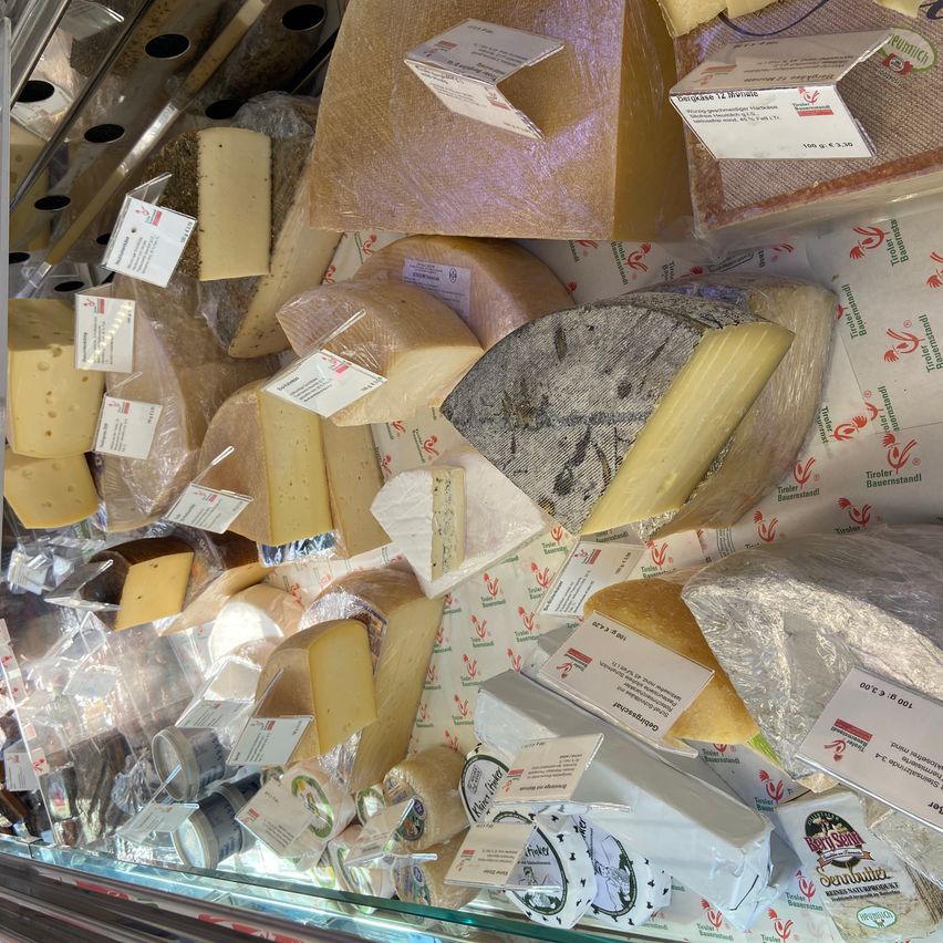 Verschiedene Käsesorten in unterschiedlichen Formen und Größen sind in einem Glasvitrinenkasten übereinander gestapelt. Jeder Käse hat ein Etikett und ist in Plastik verpackt.