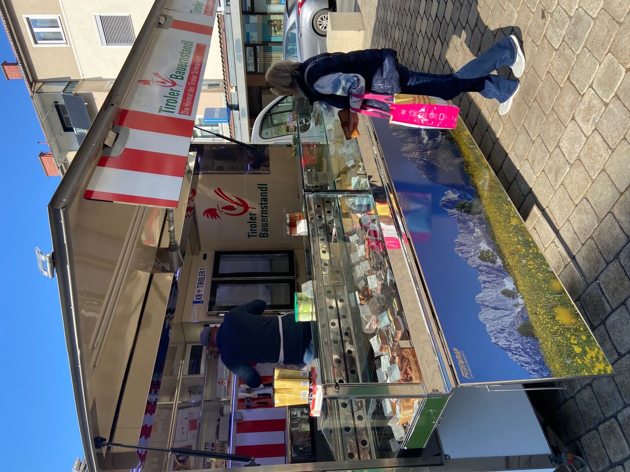 Ein Imbisswagen mit einer Vitrine und einer Frau, die einen Beutel vor einem Berglandschaftsbild hält.