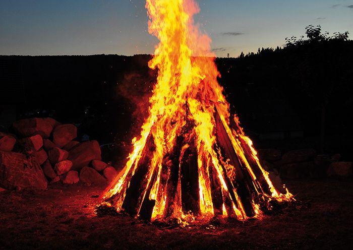 Ein Lagerfeuer brennt hell in der dunklen Nacht, umgeben von Felsen und Bäumen. Das Feuer beleuchtet das Gebiet und wirft Schatten und Wärme ab.