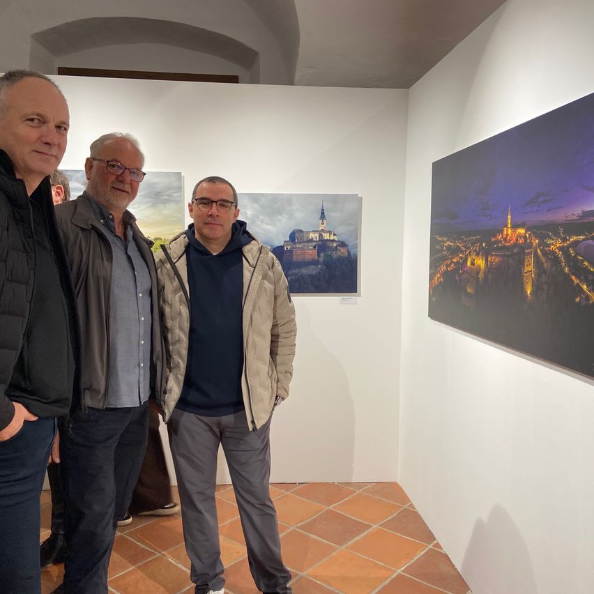 Drei Männer stehen in einer Kunstgalerie mit gefliesten Böden, tragen Jacken und Brillen. Sie stehen vor zwei großen Gemälden an der Wand, eines zeigt eine Stadt bei Nacht und das andere eine Burg in der Ferne.