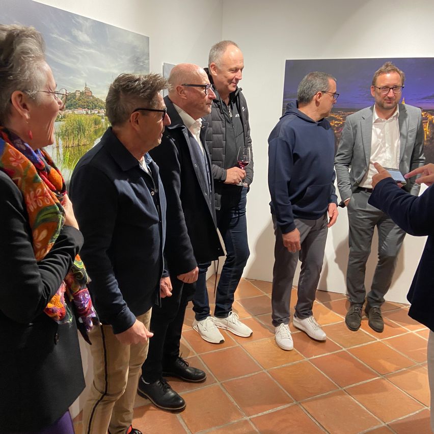 Eine Gruppe von Menschen steht in einer Kunstgalerie, einige lächeln und diskutieren die Gemälde an der Wand. Ein Mann hält ein Weinglas.