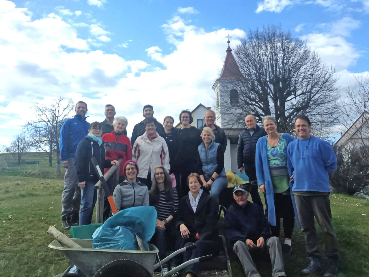 Eine Gruppe von Menschen posiert für ein Foto im Freien mit einer Kirche und einem Baum im Hintergrund, einige mit Schaufeln und andere auf Schubkarren sitzend.