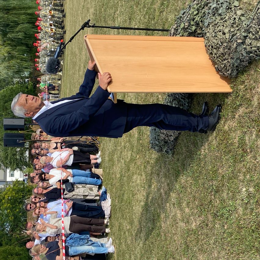 Ein Mann in Anzug steht hinter einem Rednerpult und spricht eine Menschenmenge an, die sich auf einem Rasenfeld aufstellt.