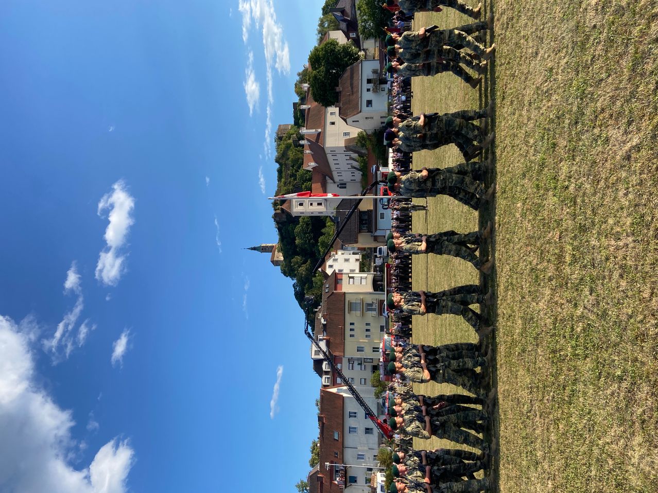 Eine Gruppe von Soldaten in Uniform steht in Formation auf einem Rasenfeld unter einem klaren blauen Himmel. Im Hintergrund sind Gebäude und Bäume zu sehen.
