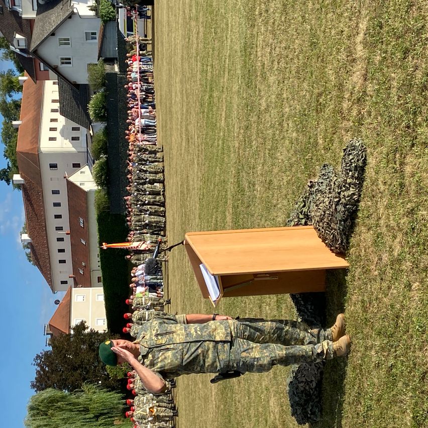 Ein Soldat salutiert vor einem Rednerpult auf einem Rasenfeld, dahinter Reihen von Soldaten, im Hintergrund Gebäude.