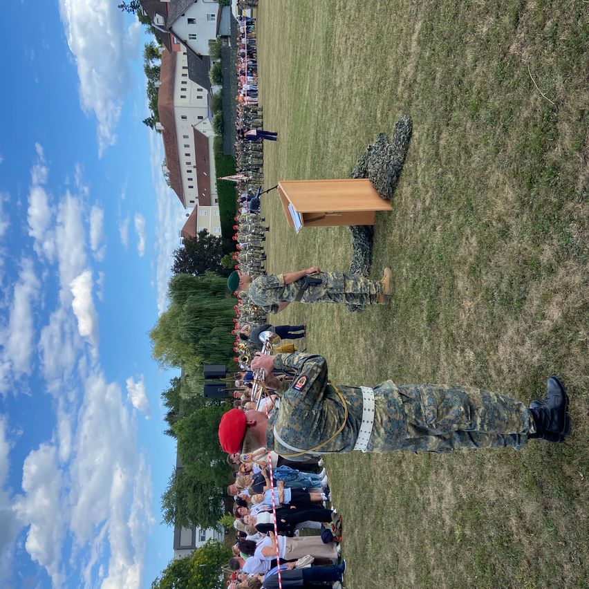 Eine militärische Zeremonie findet auf einem Rasenfeld statt, das von einer Menge Menschen beobachtet wird. Ein Soldat spielt Trompete, während ein anderer hinter einem Podium steht.