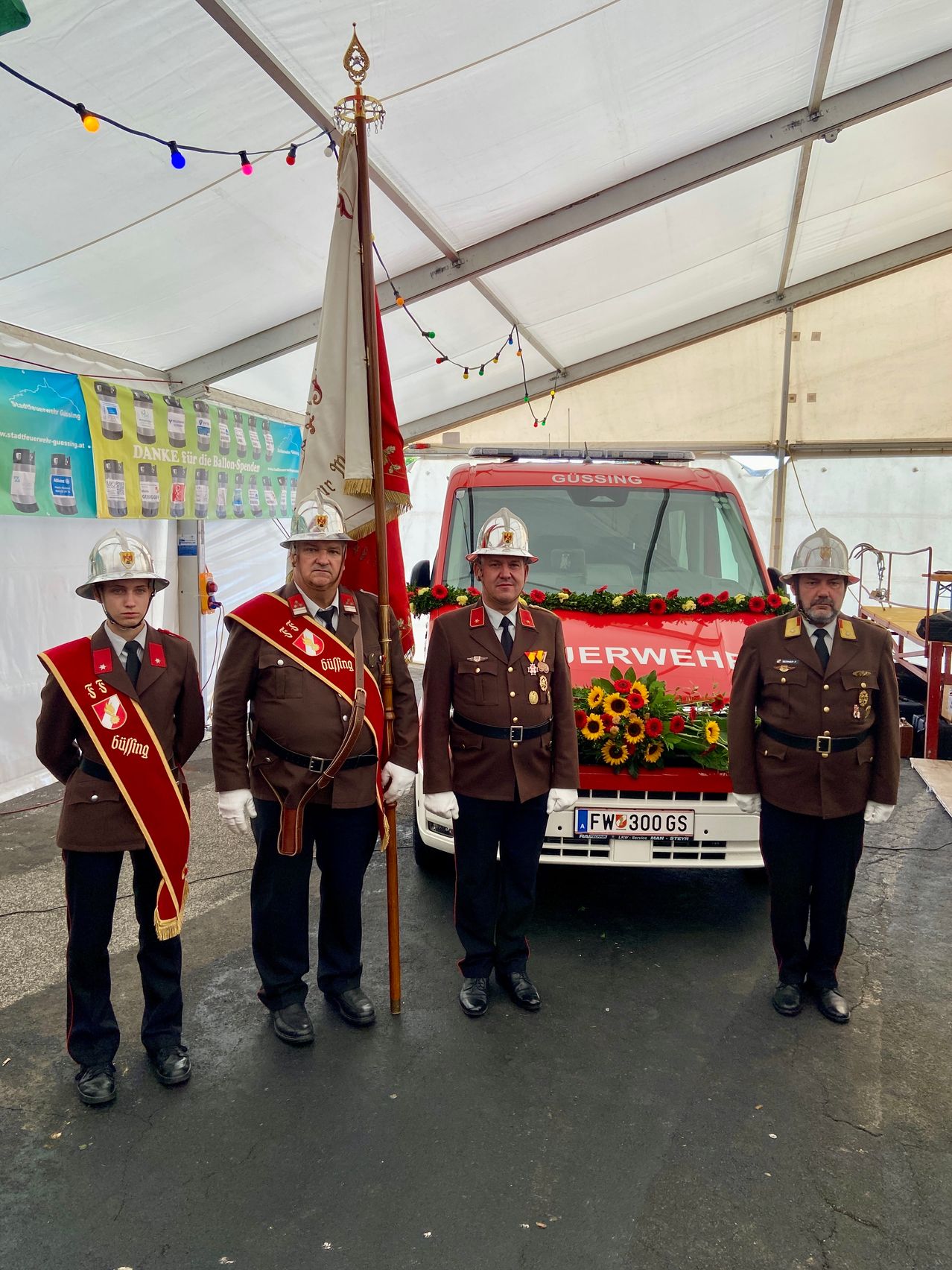 Vier Feuerwehrleute in Uniform stehen vor einem roten Feuerwehrauto mit Blumen auf der Motorhaube. Sie sind unter einem Zelt mit einem Banner im Hintergrund.