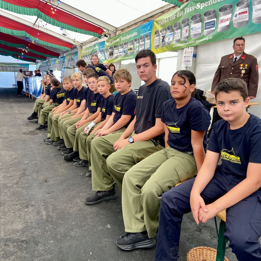 Eine Gruppe junger Jungen in Uniform sitzt auf einer Bank in einem Zelt, mit einem Militäroffizier hinter ihnen.