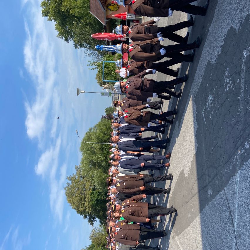 Eine Gruppe uniformierter Personen, möglicherweise Militärangehörige, steht in einer Reihe auf einer Straße unter einem blauen Himmel mit verstreuten Wolken.
