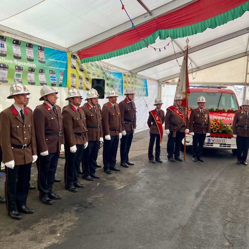 Eine Gruppe von Feuerwehrleuten in Uniform steht in einer Reihe unter einem Zelt mit rotem und grünem Vordach, in der Nähe eines Feuerwehrwagens.