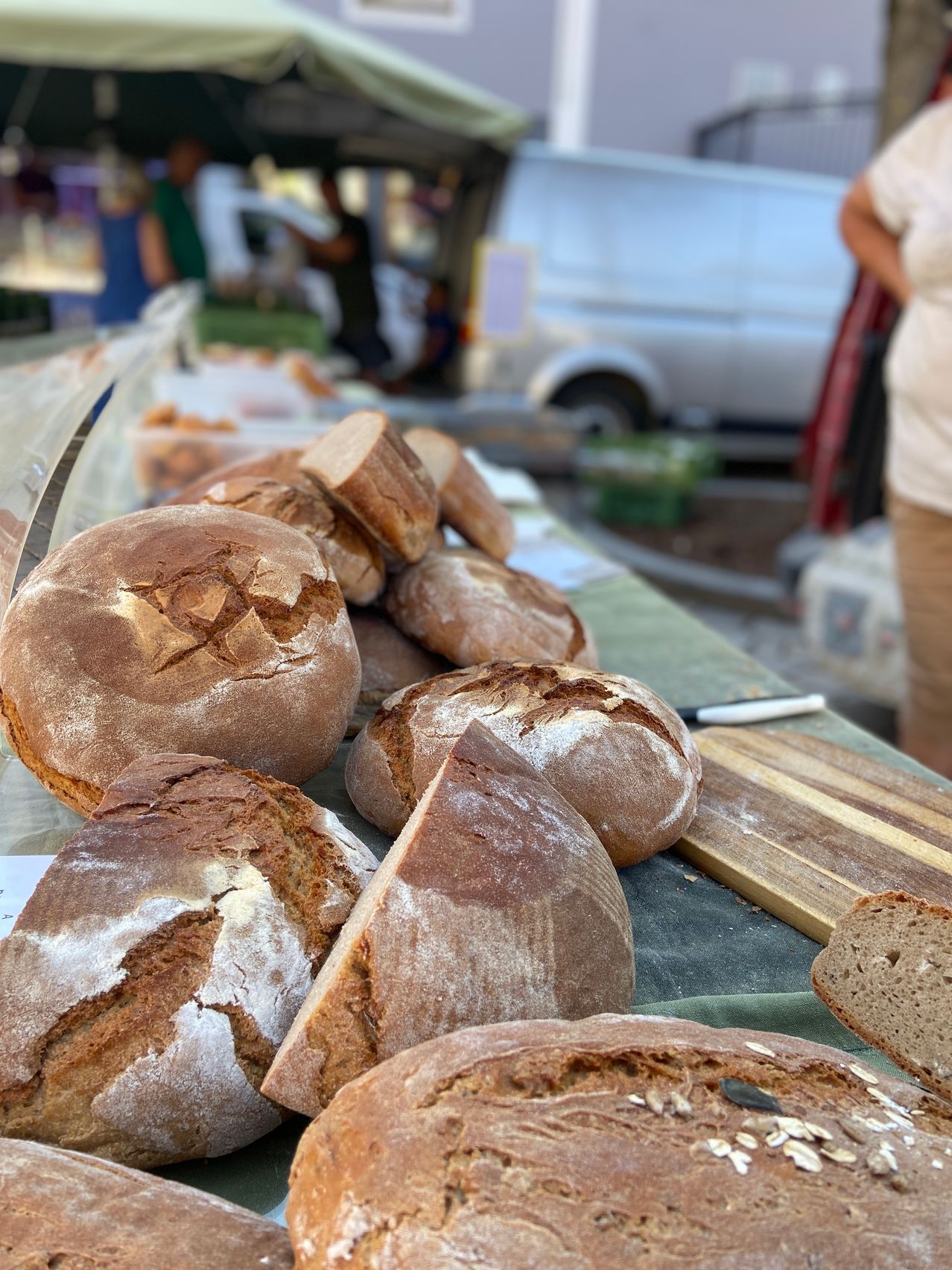 Ein Tisch voller handwerklicher Brote mit verschiedenen Schnitten und Formen, ausgestellt auf einem Outdoor-Markt mit einem Lieferwagen und Menschen im Hintergrund.
