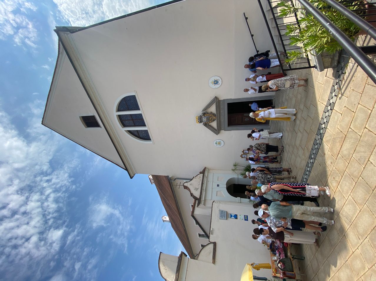 Eine Gruppe von Menschen versammelt sich vor einer Kirche, einige stehen in der Nähe des Eingangs, während andere weiter entfernt sind. Die Kirche hat eine weiße Außenfassade mit dekorativen Elementen und Fenstern.
