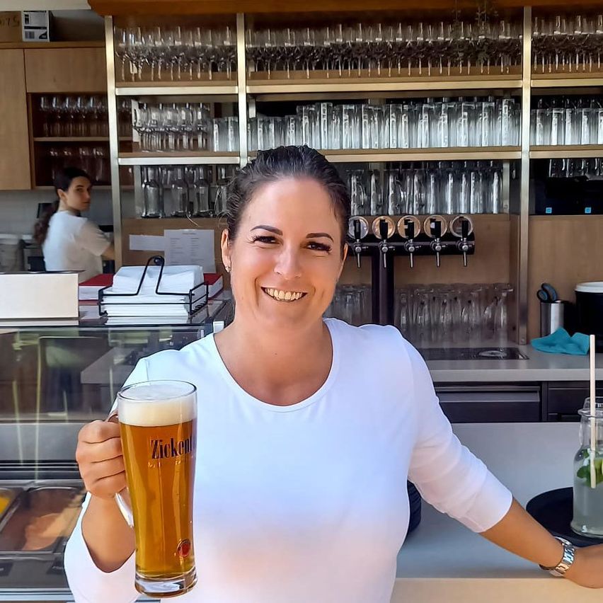 Eine Frau in einem weißen T-Shirt hält ein volles Glas Bier in einer Bar. Hinter ihr sind Regale mit Gläsern und Flaschen zu sehen. Eine weitere Person steht im Hintergrund.