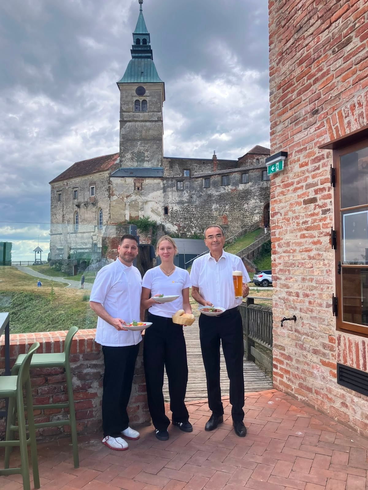 Drei Personen in weißen Kochuniformen stehen vor einem Backsteingebäude, halten Teller mit Essen und ein Glas Bier. Dahinter ist ein Schloss mit einem Turm gegen einen bewölkten Himmel sichtbar.