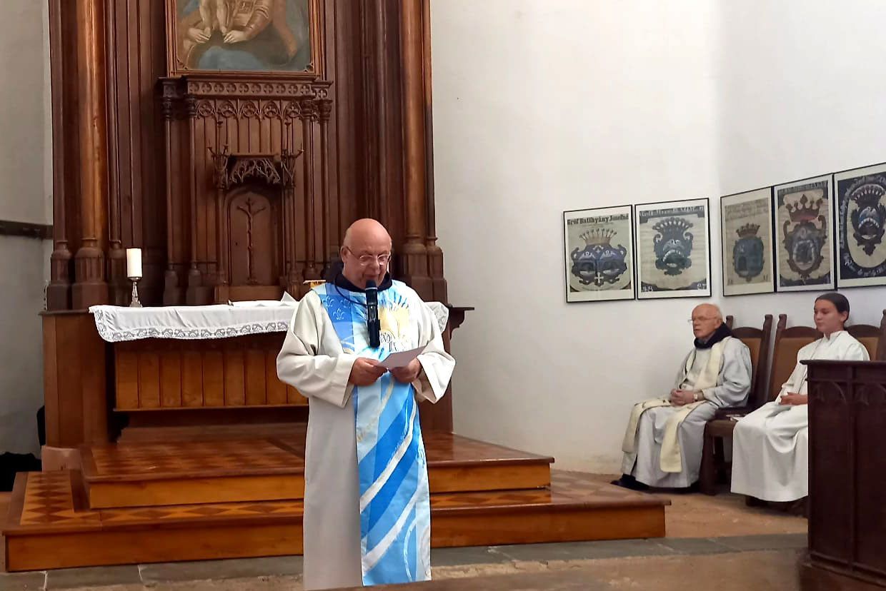 Ein Priester steht vor einem hölzernen Altar, hält ein Papier und ein Mikrofon in der Hand, mit einem anderen Priester, der auf einer Bank hinter ihm sitzt.