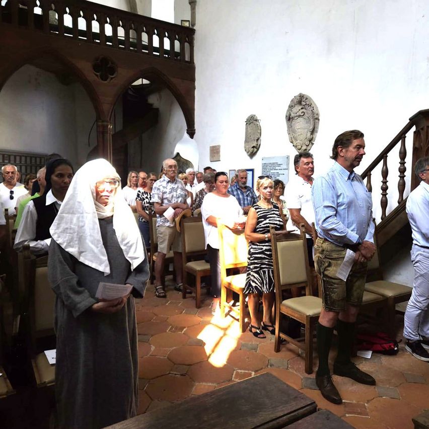 Eine Gruppe von Menschen steht in einer Kirche, einige in traditioneller Kleidung, während andere lässig gekleidet sind. Eine Frau mit einem weißen Kopftuch hält ein Stück Papier.