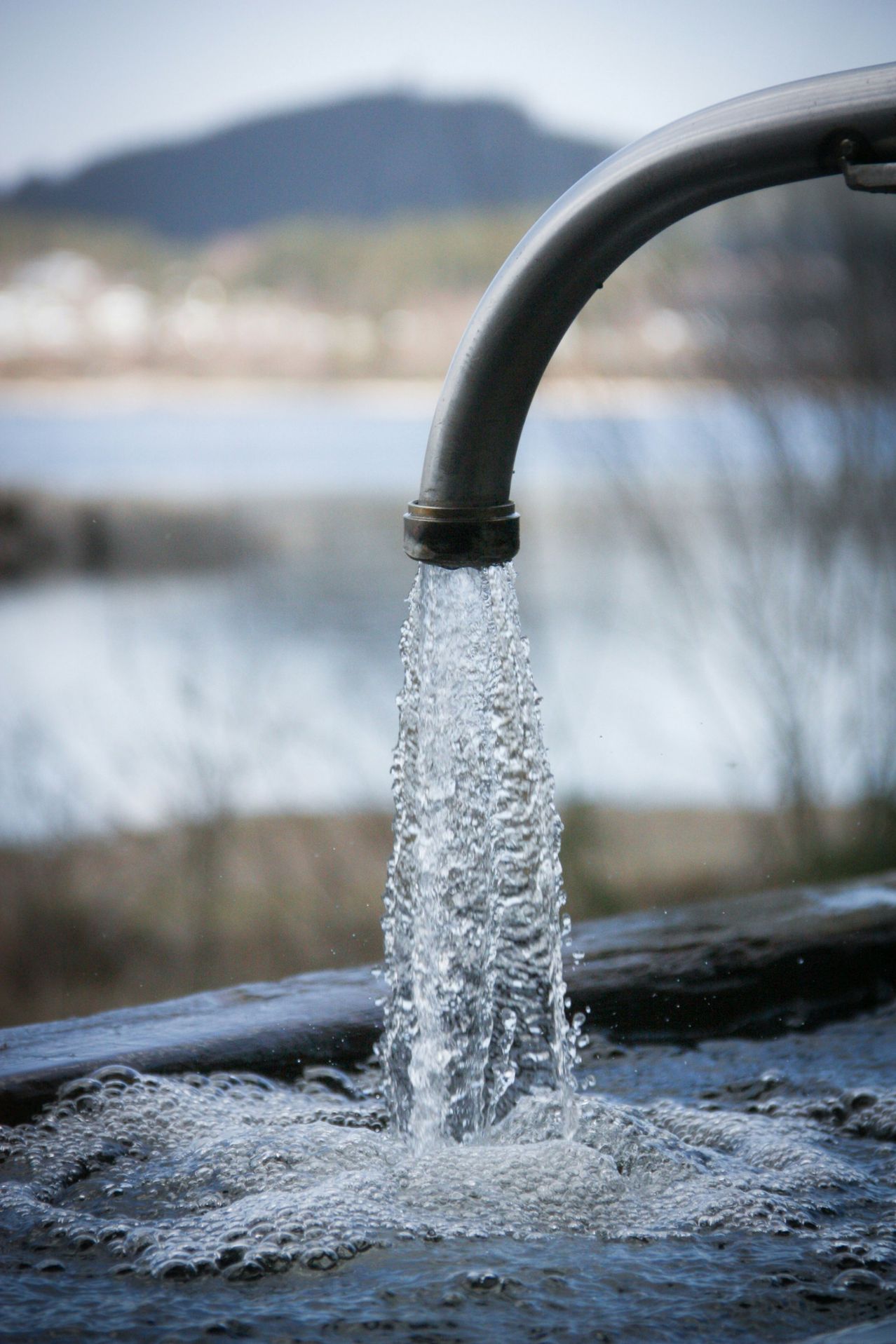 Wasser fließt aus einer Metallrohrleitung in einen Pool, mit einem See und verschwommenem Hintergrund.