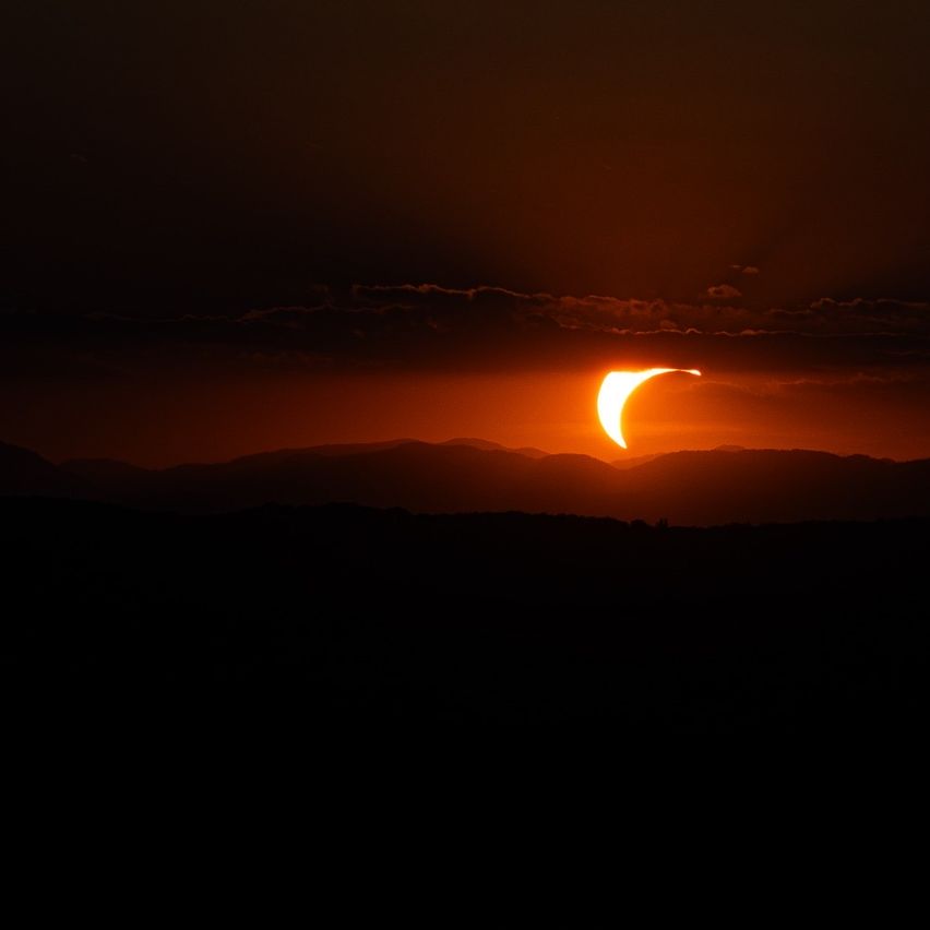 A partial solar eclipse is occurring at sunset with the sun partially obscured by the moon. The sky is orange and dark, with mountains in the background.