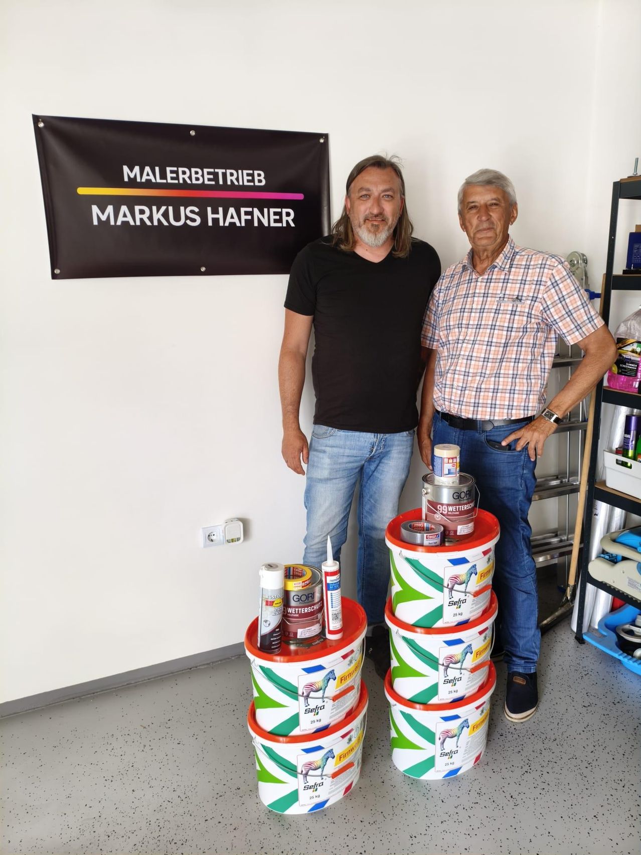Two men stand in a room with paint cans and materials. A banner reads Malerbetrieb Markus Hafner.