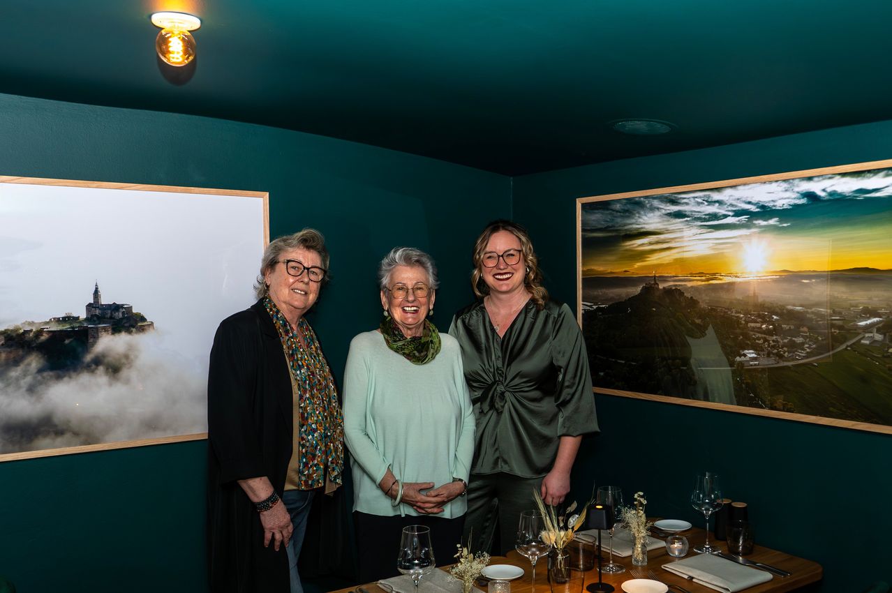 Three women stand together in a room, smiling and posing for a photo. They are surrounded by a table set for a meal.
