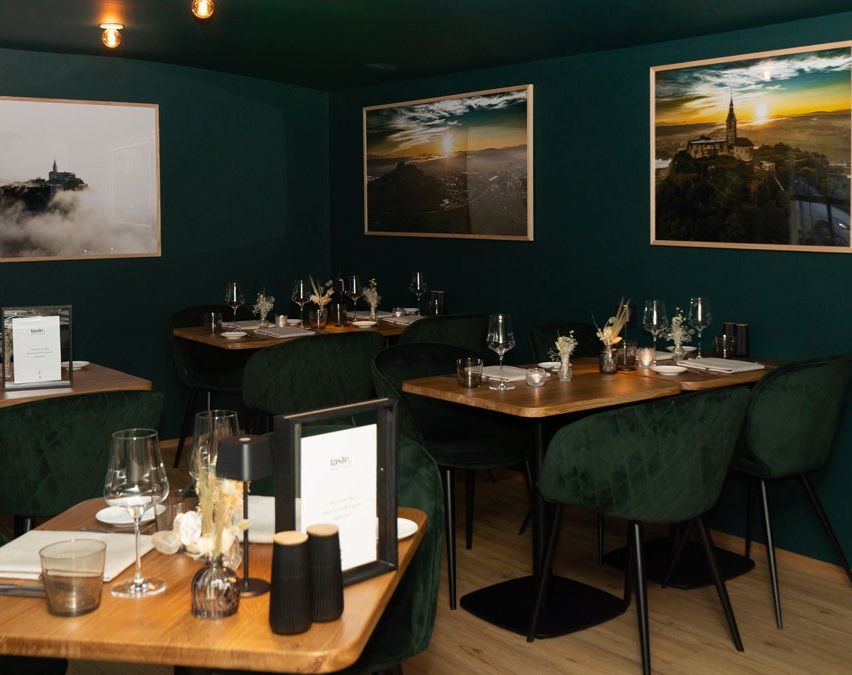 A restaurant with a wooden table and green chairs. The table has glasses, plates, and other items. The room has green walls and three frames on the wall. The room is illuminated with warm lights.