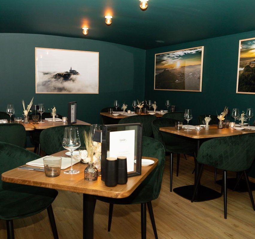 A restaurant with green walls and wooden floors. It has tables and chairs, each with wine glasses, plates, and menus. There are also picture frames on the walls.