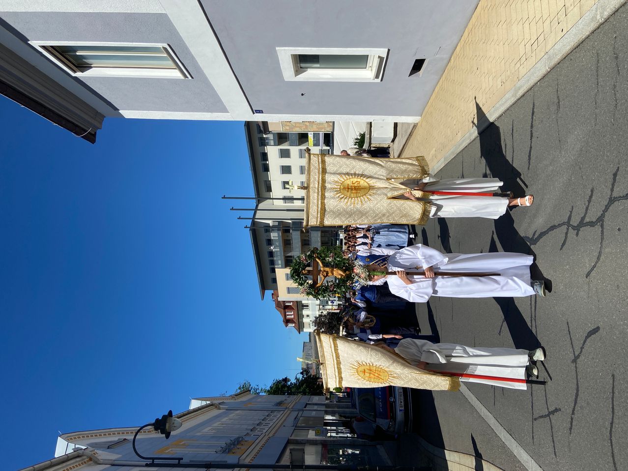 A religious procession is underway on a sunny day in a city. The clergy are carrying banners with sun symbols. The buildings and streetlights are visible in the background.