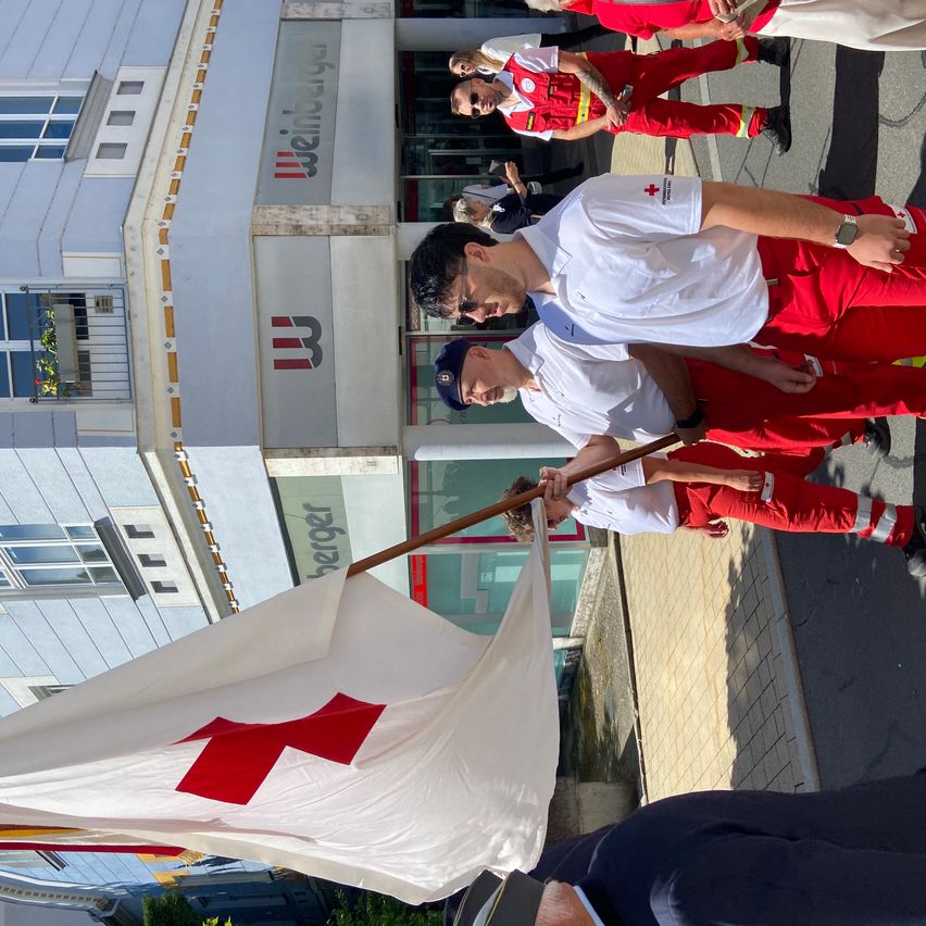 Red Cross volunteers are marching in a parade outside a building with the sign 'Weinberger'. They are holding a flag and wearing white and red uniforms.