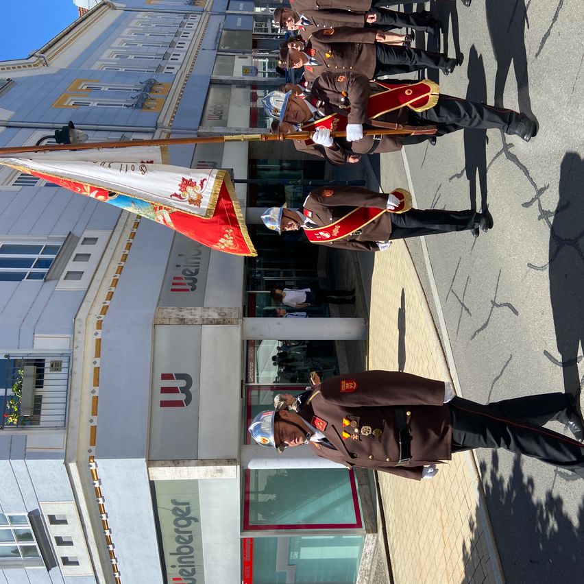 Soldiers in uniform stand in a row, holding flags and batons, in front of a building. A person is visible in the background near the entrance.