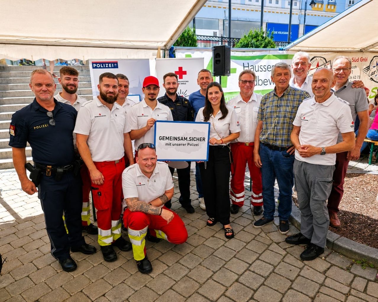 Eine Gruppe von Menschen, einige in Uniform, steht auf einer Ziegelfläche unter einem Zelt. Eine Frau hält ein großes weißes Schild. Hinter ihnen sind Banner mit Logos.