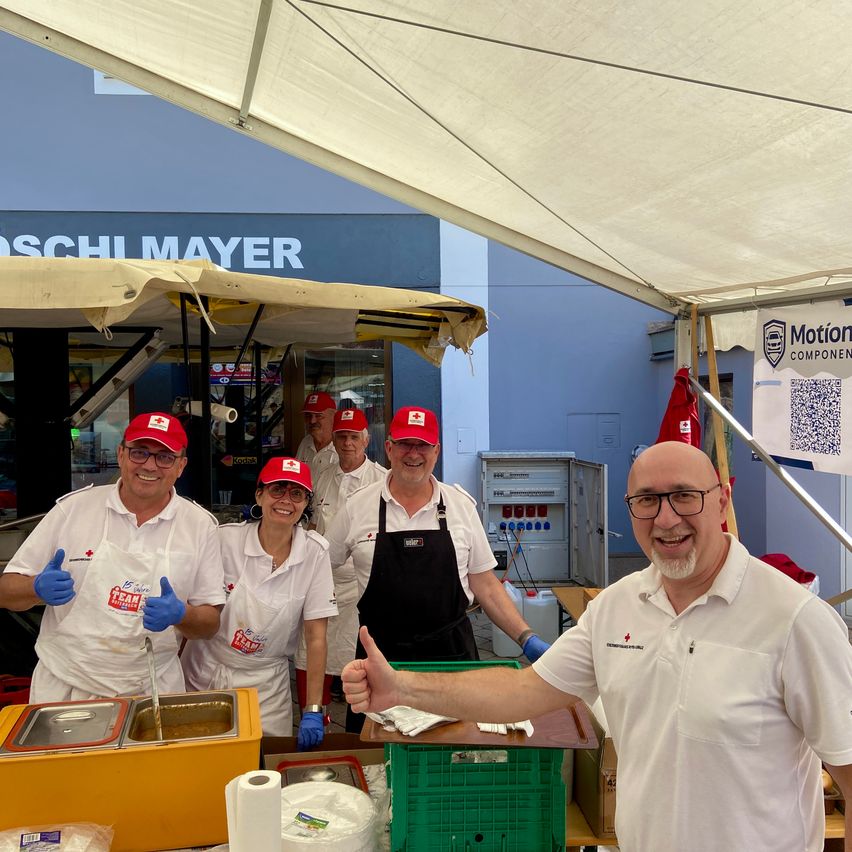 Eine Gruppe von Menschen in roten Hüten und weißen Schürzen, möglicherweise Freiwillige, steht unter einem Zelt. Ein Mann in einem weißen Hemd und Brille zeigt den Daumen nach oben. Vor ihnen befinden sich Essensbehälter und ein Tisch. Dahinter ist ein Gebäude mit einem Schild zu sehen, auf dem 'Schili Mayer' steht.