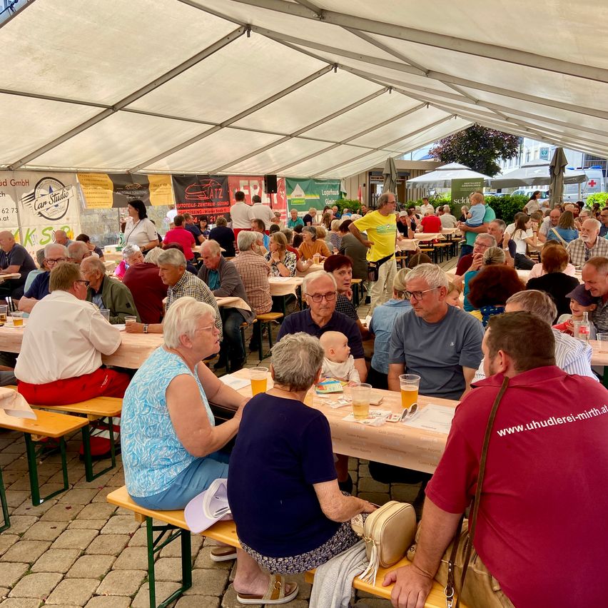 Viele Menschen sitzen unter einem Zelt an Tischen, einige essen und trinken, während andere in Gespräche vertieft sind. Im Hintergrund sind Banner zu sehen.