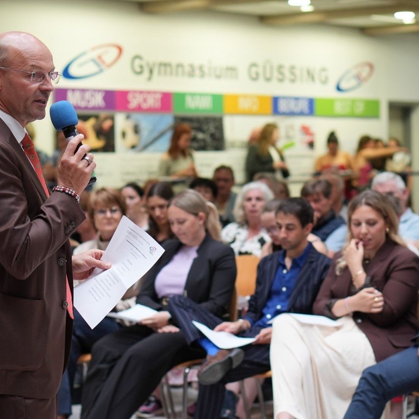 Ein älterer Mann in Anzug hält ein Mikrofon und liest von einem Papier im Gymnasium Gussing. Verschiedene Personen sitzen auf Stühlen und hören aufmerksam zu.