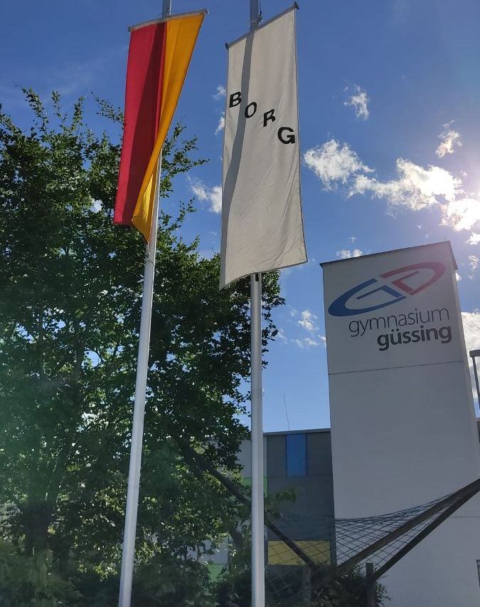 Ein Fahnenmast mit einer deutschen Flagge und einer weißen Flagge mit 'BORG' steht vor dem Gussing Gymnasium-Gebäude unter einem blauen Himmel mit Wolken.