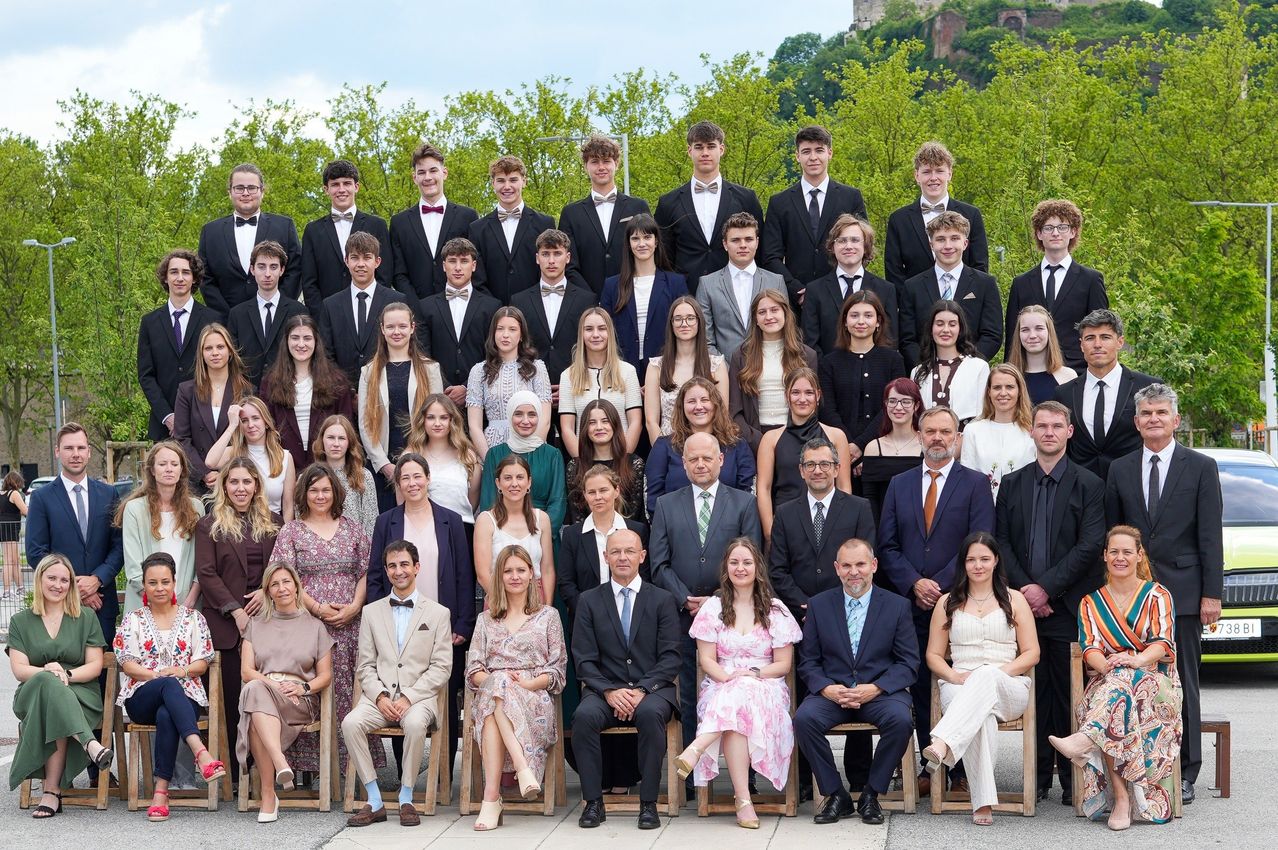 Eine große Gruppe von Schülern und Lehrern in formeller Kleidung posiert für ein Foto, einige sitzend und andere stehend, in einem Park mit Bäumen und einer Burg im Hintergrund.