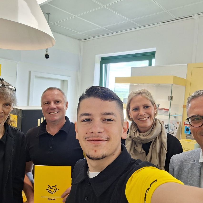 Fünf Personen lächeln für ein Foto. Ein Mann hält ein gelbes Buch mit einem Händeschüttelbild und dem Wort Danke. Hinter ihm hält ein Mann eine gelbe Box. Eine Frau trägt eine Brille. Der Raum hat eine Lampe, ein Fenster und einen gelben Schrank.
