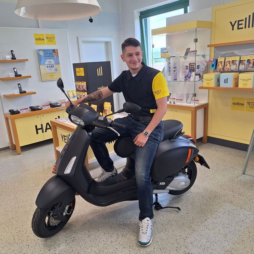 Ein Mann in einem schwarz-gelben Shirt sitzt auf einem schwarzen Roller in einem Geschäft. Er lächelt und posiert für ein Foto. Hinter ihm ist eine gelbe Wand mit einem Poster.