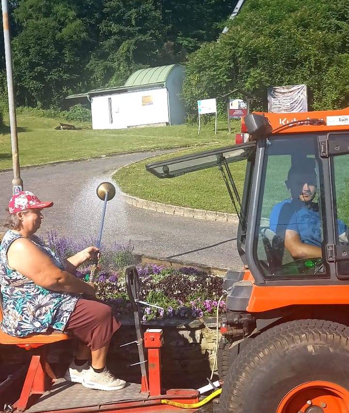 Eine Frau in einer Mütze sitzt auf einer Bank und hält eine Gießkanne in der Hand, neben einem Traktor. Ein Mann sitzt im Traktor, und der Traktor ist auf einer Straße geparkt.