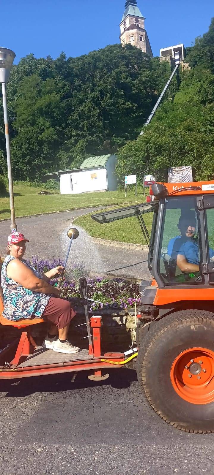 Eine Frau mit einer Mütze fährt einen orangefarbenen Traktor und sprüht Wasser in einem Garten. Ein Mann sitzt im Inneren des Traktors und lächelt. Der Traktor ist neben einem Rasen abgestellt.