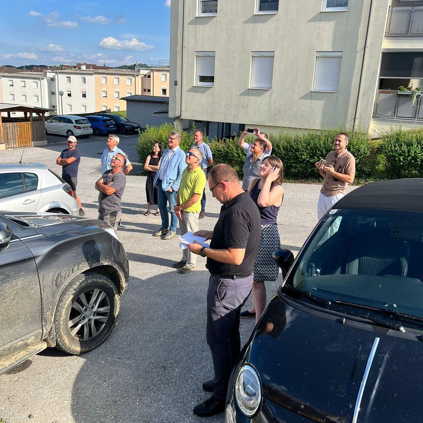 Eine Gruppe von Menschen ist um zwei Autos in einem Parkplatz versammelt. Ein Mann hält ein Papier und spricht zu ihnen. Der Parkplatz befindet sich in der Nähe eines Wohnhauses.